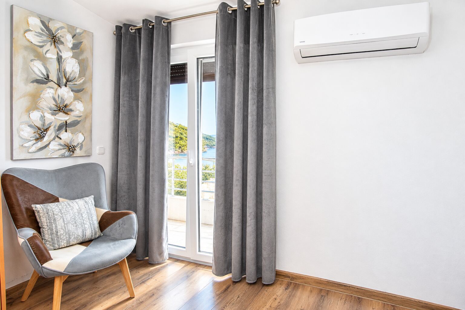 A room with a grey chair, grey curtains, a painting of flowers, and an air conditioning unit.
