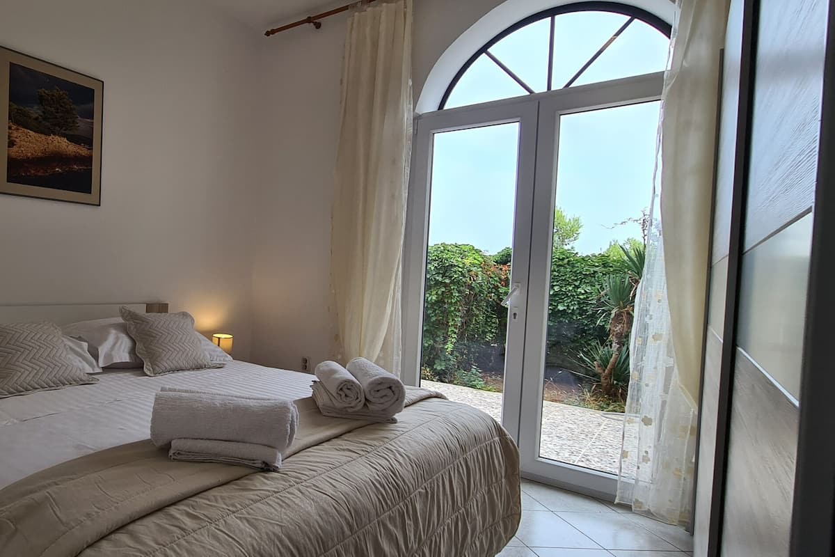 Un dormitorio con una cama grande, una mesita de noche, una lámpara, un cuadro y vistas a través de una ventana.