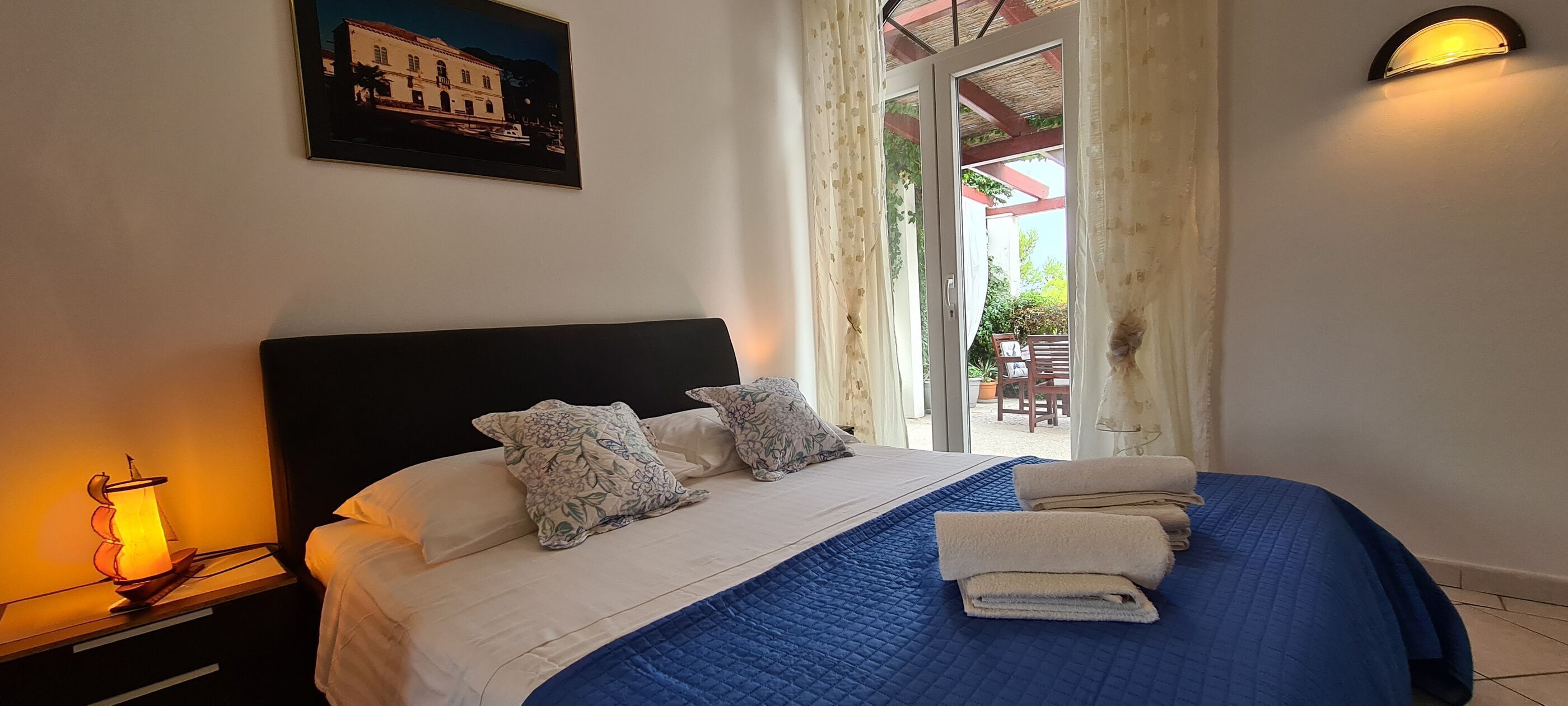A bedroom with a bed, pillows, a nightstand, a lamp, a painting, and a view of the outside.