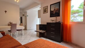 A modern living room with a sofa, a dining table with chairs, a TV, and a cabinet. There is a window with curtains, and artwork on the walls. - Apartment Nikola (Jelsa)