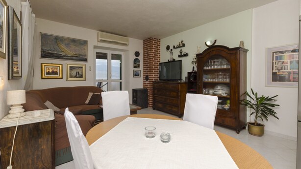 A living room with a sofa, a dining table with two glasses, a TV, a brick fireplace, and a wooden cabinet with glassware. - Apartments Jurica (Postira)