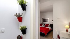 Un couloir agrémenté de plantes en pot, un lit avec une literie rouge, un bureau avec une lampe et une chaise.