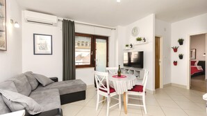 Un salon moderne avec un canapé gris, une table à manger avec des chaises blanches, un téléviseur à écran plat et une grande fenêtre avec des rideaux.