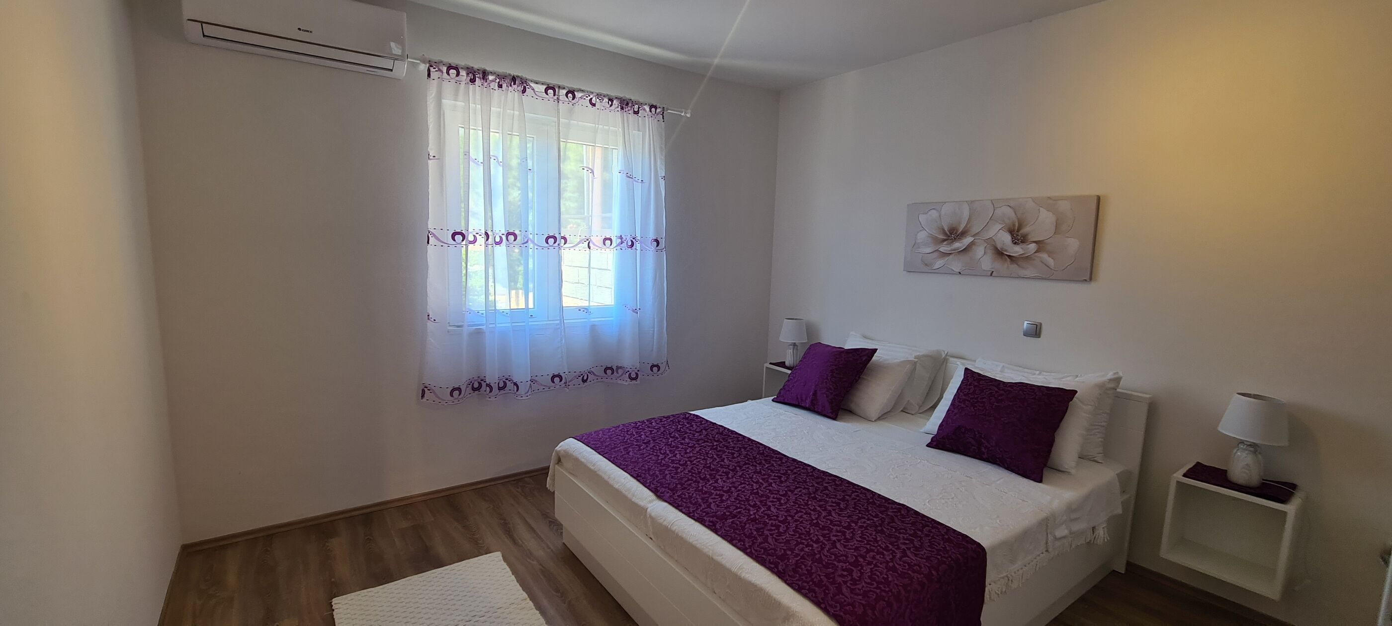 A bedroom with a bed, a window with sheer curtains, a wall-mounted artwork, and a bedside table with a lamp.