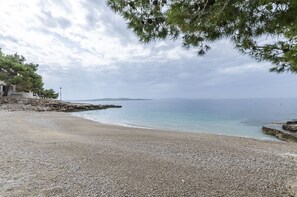 Une plage de galets bordée d’un muret et d’arbres, qui s’étend jusqu’à une mer calme et des collines lointaines.