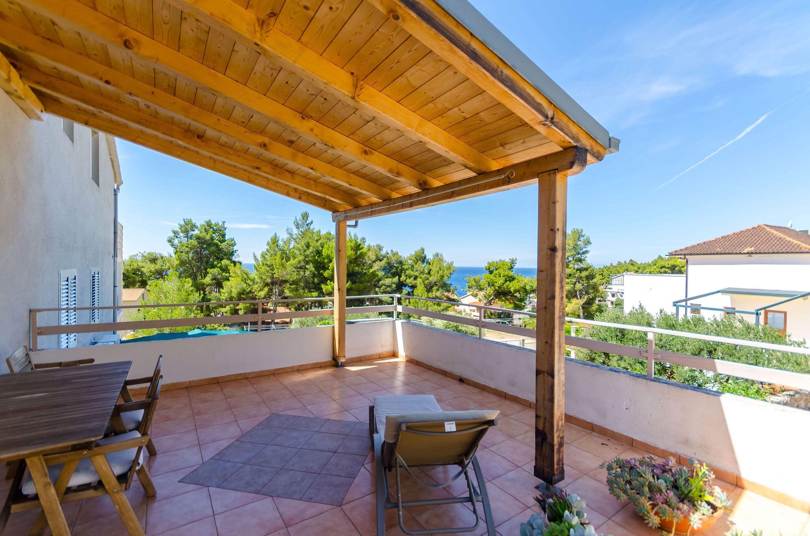 Eine überdachte Terrasse mit Holzboden, einem Tisch, Stühlen und Blick aufs Meer.