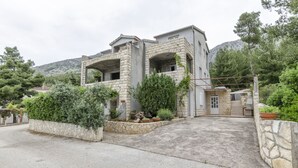 Une maison en pierre dotée d’un balcon, entourée de verdure et avec une chaîne de montagnes en arrière-plan.