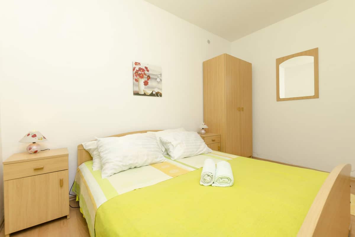 A neatly made bed with a green bedspread and two pillows, a wooden nightstand with a lamp, a wooden wardrobe, and a wall-mounted mirror.
