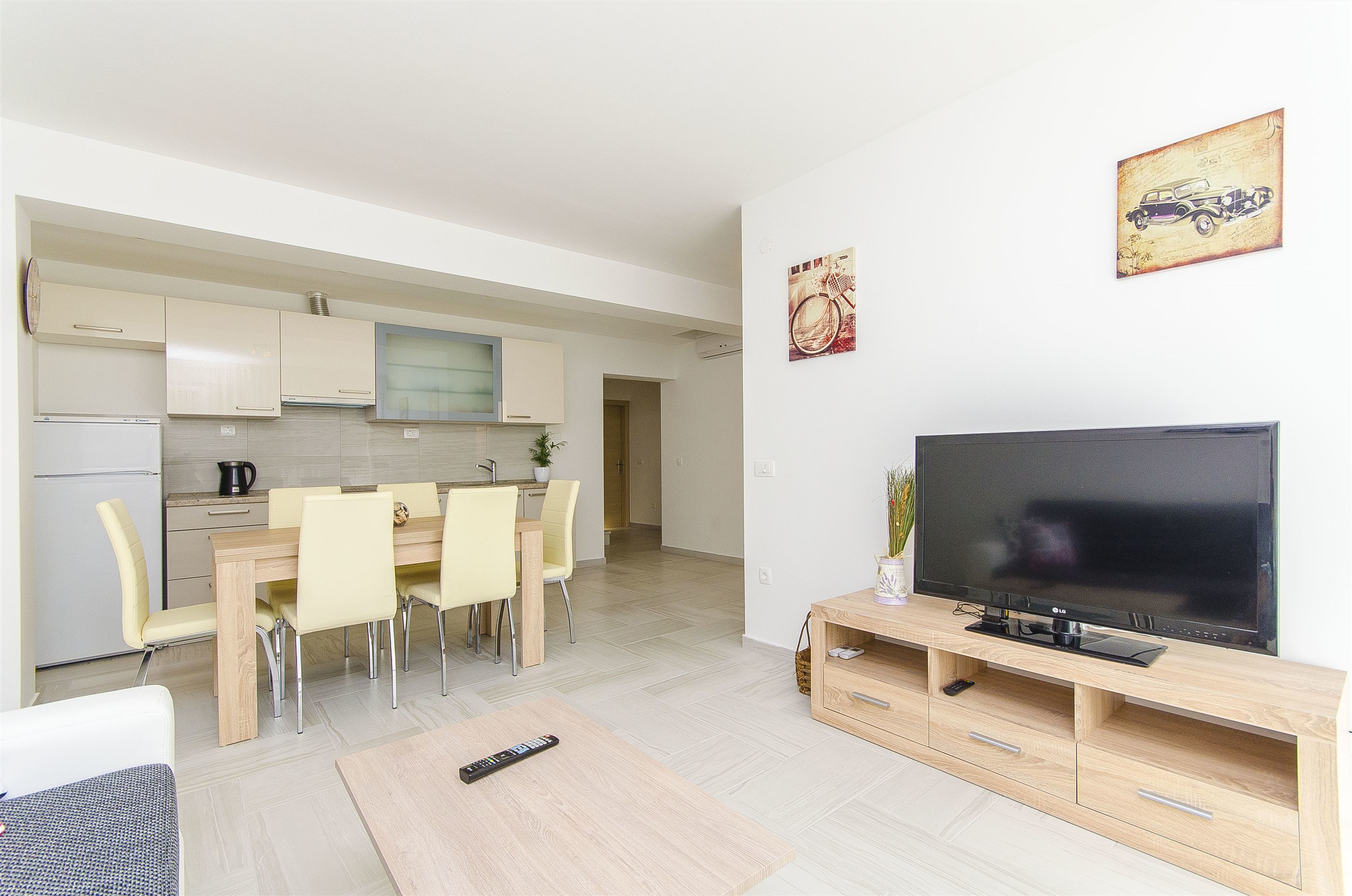 Une cuisine moderne avec des meubles blancs, un coin repas avec une table et des chaises en bois, et un salon avec un téléviseur posé sur un meuble en bois.