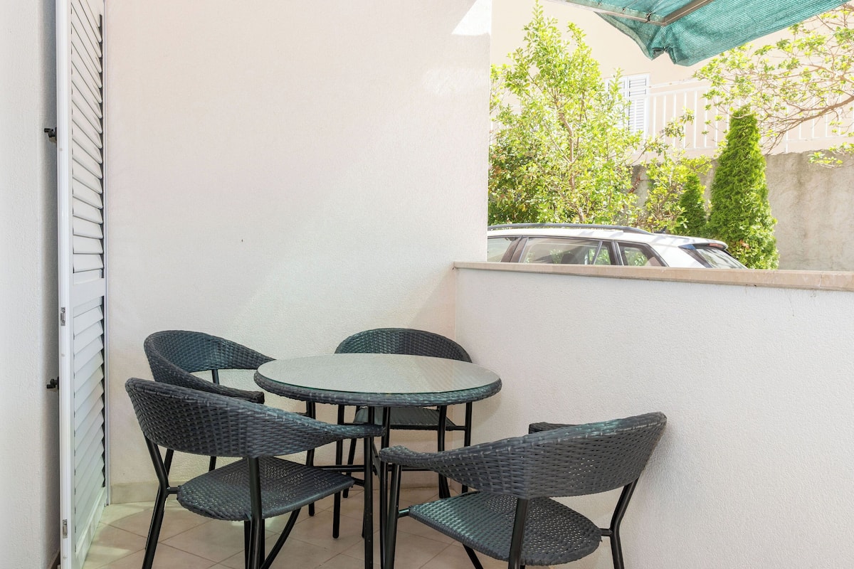 A balcony with a round table and four chairs, a car parked outside, and a view of trees and a building in the background.