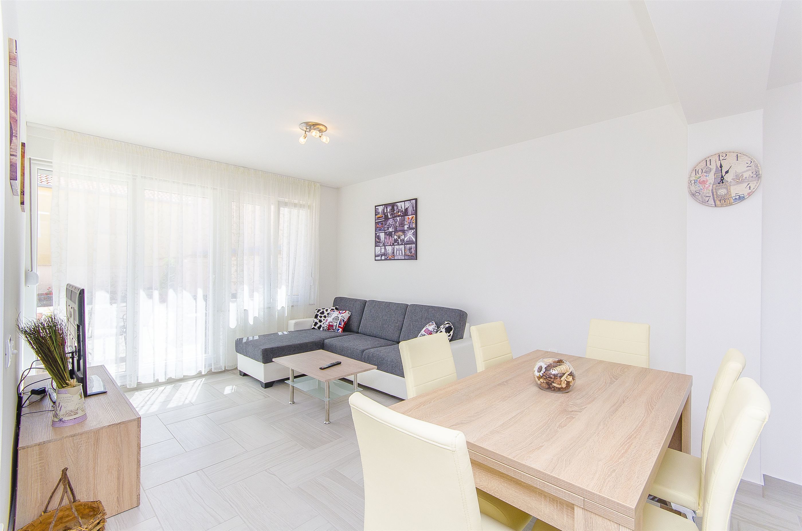 A modern living room with a sofa, dining table, and chairs. There is a clock on the wall and a picture frame.