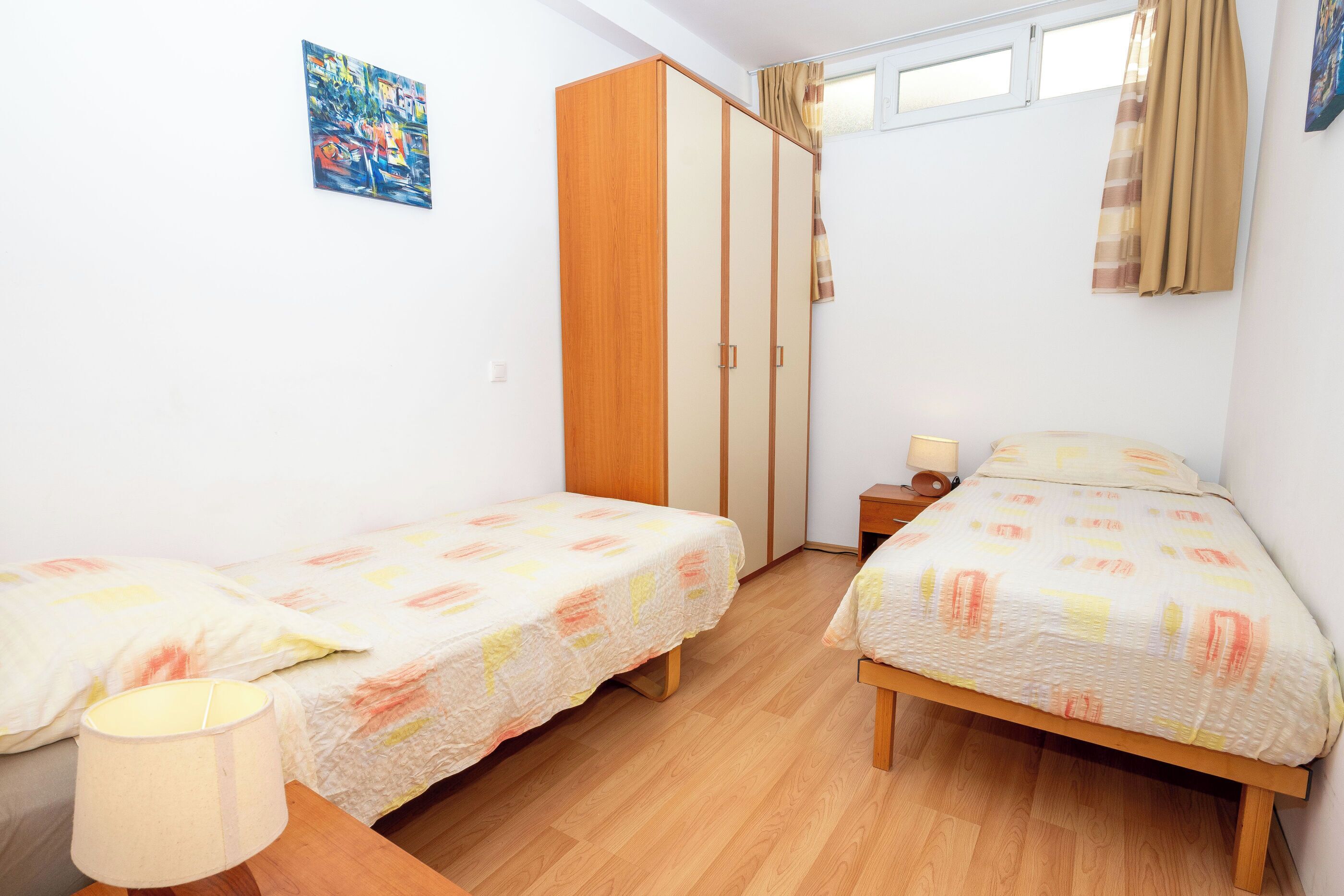 A bedroom with two single beds, a wardrobe, a nightstand, a lamp, and a window with curtains.