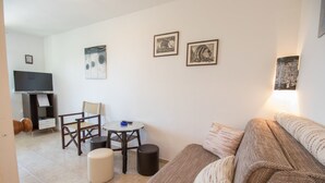 Ein Wohnzimmer mit einer Couch, einem Fernseher, einem kleinen Tisch und zwei Stühlen. An der Wand hängen gerahmte Bilder und rechts steht eine Lampe.