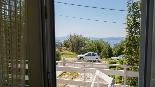 Ein Balkon mit weißer Brüstung, einer Bank und Blick auf ein Auto und Grünflächen.