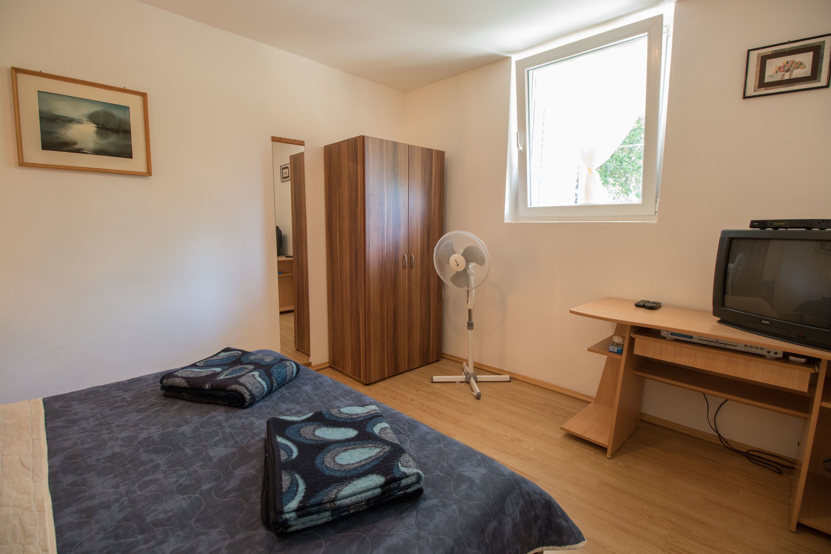 A bedroom with a bed, a wardrobe, a desk with a TV, a standing fan, and a window.