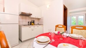 A compact kitchen with white cabinets, a red dining table set with plates and glasses, and a window with a view of greenery. - Apartments Bogdan (Pisak)