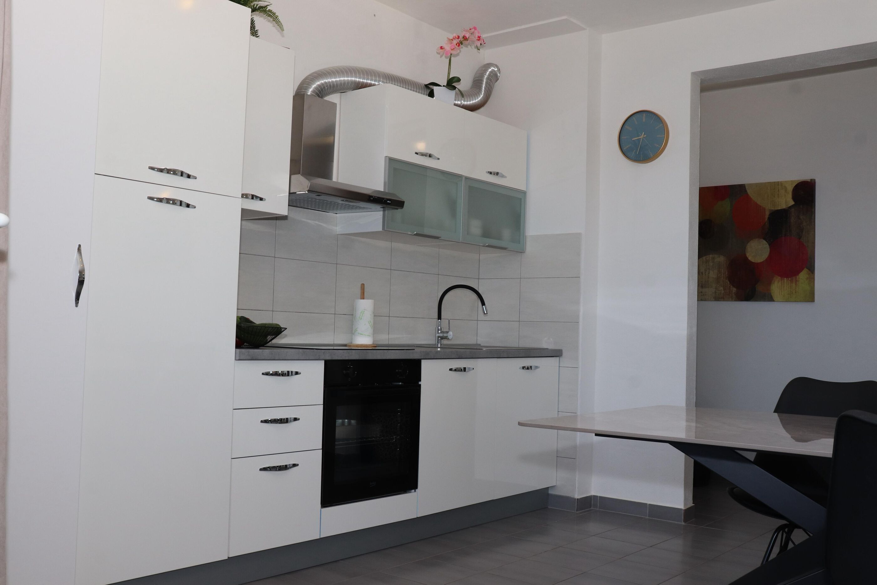 A modern kitchen with white cabinets, a stainless steel range hood, a black dishwasher, and a dining area with a table and chairs.