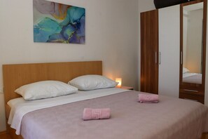 Quarto com cabeceira de madeira, cama com roupa de cama branca e rosa, toalha rosa na cama, mesa de cabeceira com abajur e guarda-roupa de madeira.