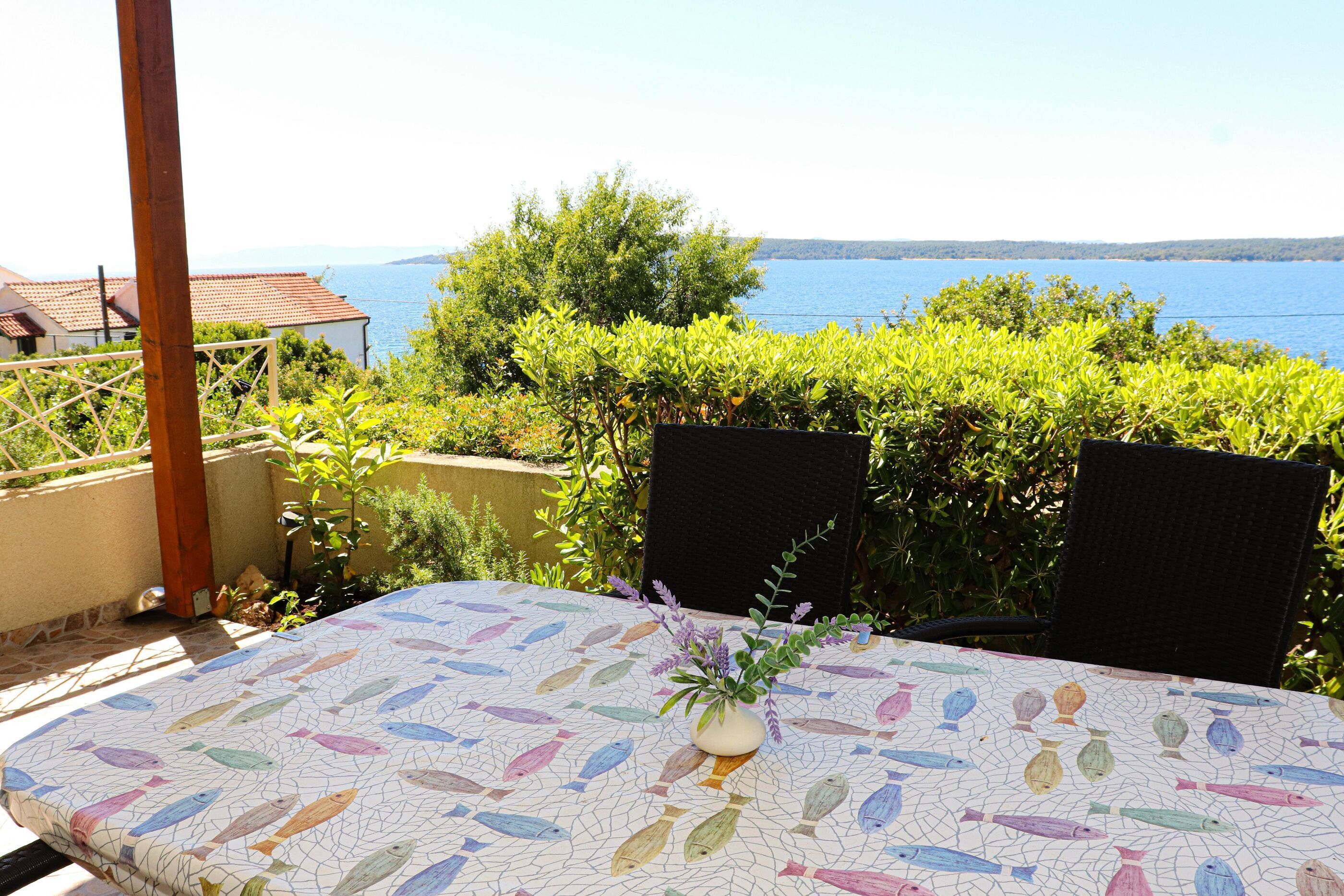 A table with a fish pattern tablecloth, a vase with flowers, and a view of the sea.