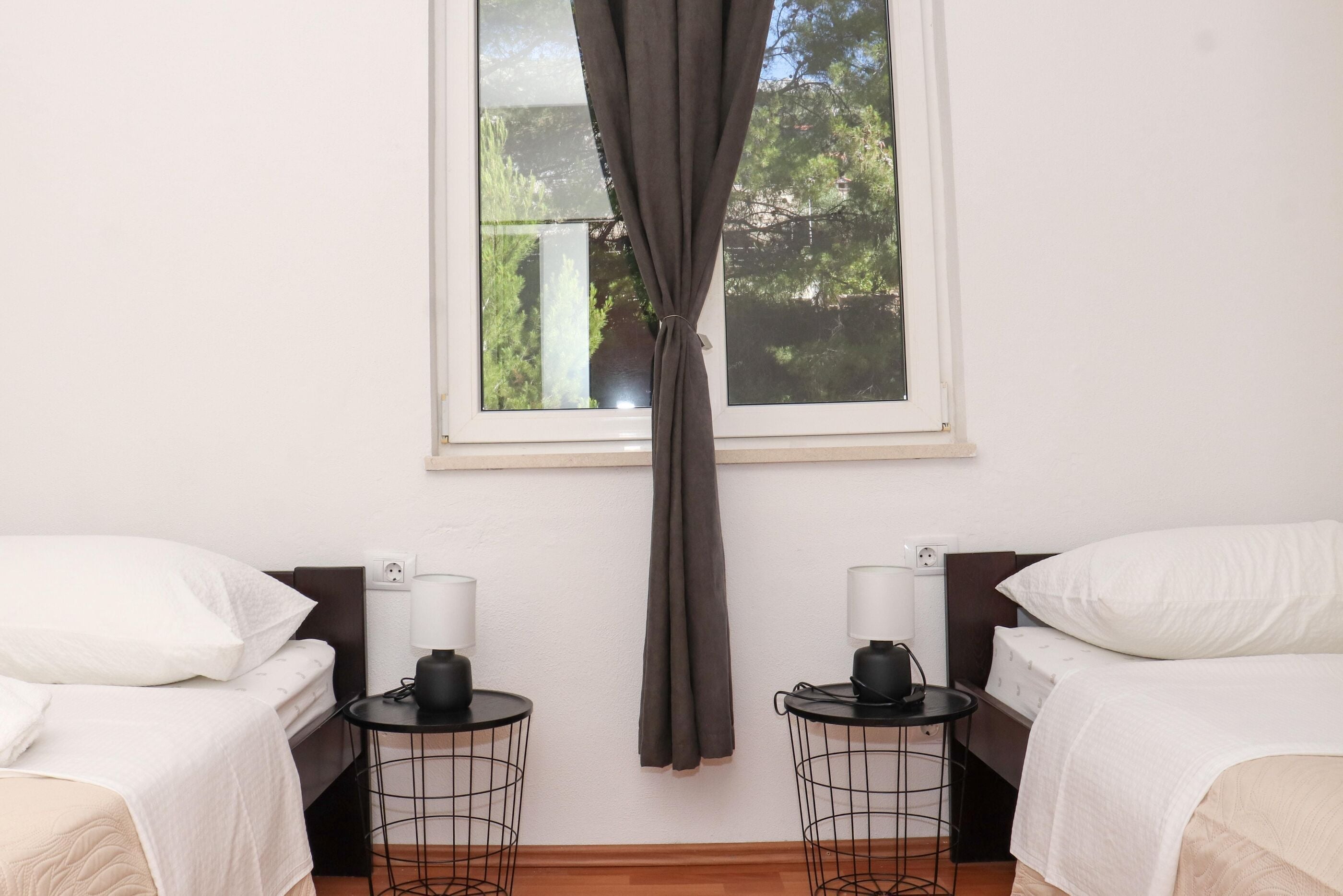 Ein Zimmer mit zwei Betten, einem fensterbeprägten Ausblick auf Grünflächen, zwei Nachttischen mit Lampen und Vorhängen.
