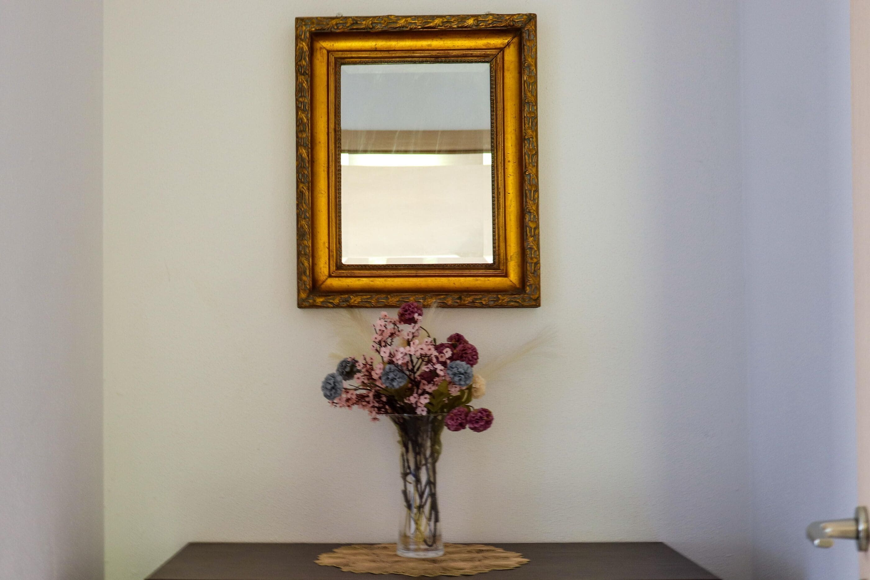 Un jarrón con flores sobre una mesa, un espejo en la pared y una puerta a la derecha.