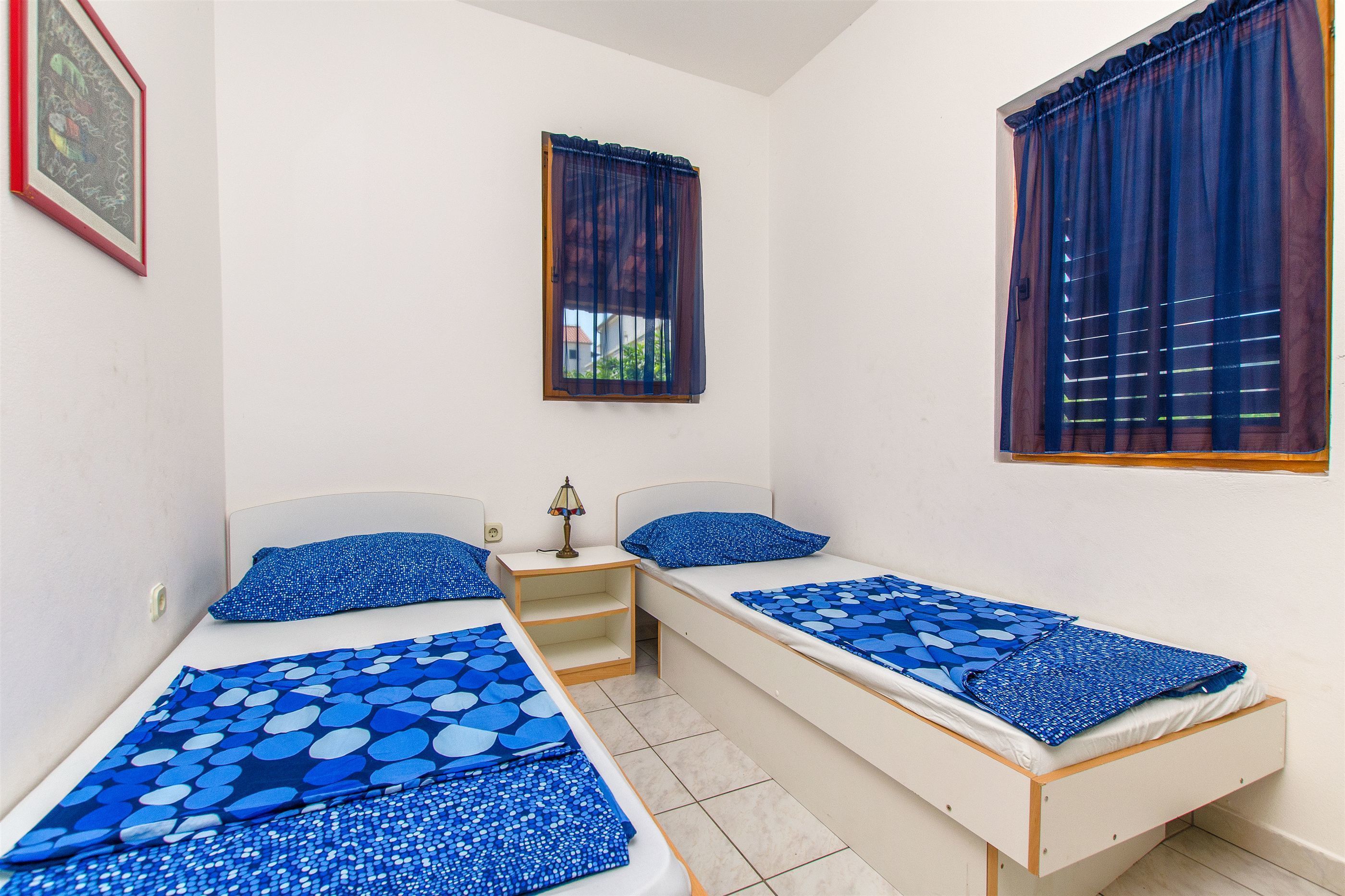 Habitació amb dos llits individuals, cadascun amb roba de llit i almoadines blaus, una petita taula de nit amb una làmpada, i una finestra amb cortines blaves.