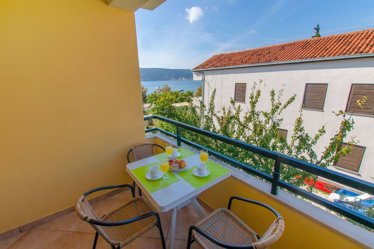 A balcony with wicker chairs, a table with breakfast, and a view of the sea and buildings.