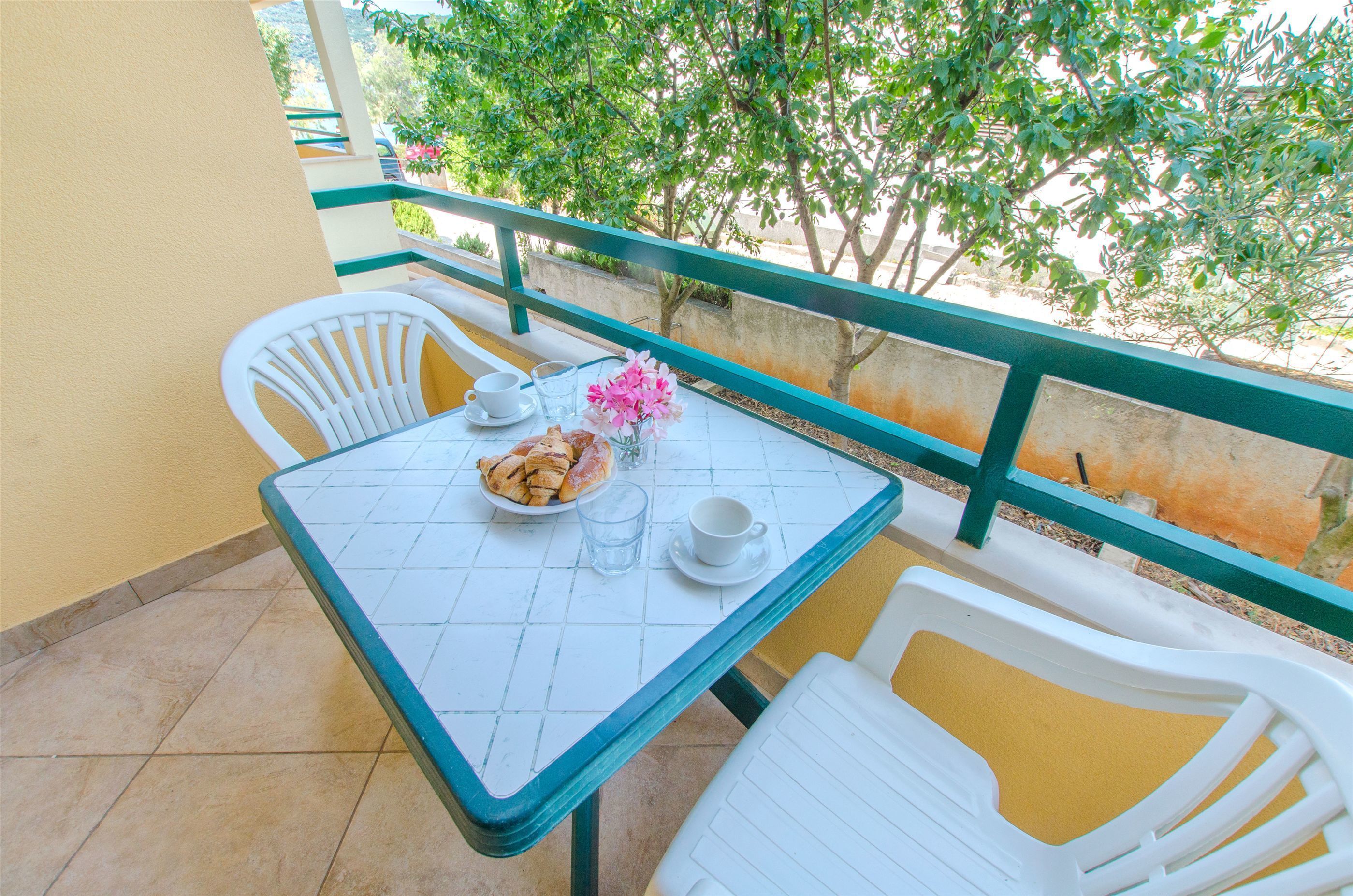 A small outdoor balcony with a table set for two, including a plate of pastries, a glass of water, and a cup.