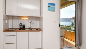Una cucina compatta con un piano di lavoro in legno, un lavello e una macchina da caffè. Una porta finestra dà accesso a un balcone con vista su un corpo d'acqua e un paesaggio lontano.