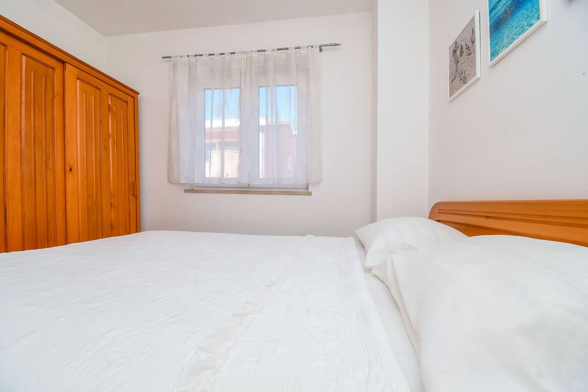 A bedroom with a wooden wardrobe, a bed with white bedding, a window with sheer curtains, and a framed picture on the wall.