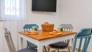 Un'area pranzo con un tavolo in legno, quattro sedie, un televisore fissato al muro e una finestra con tende leggere.