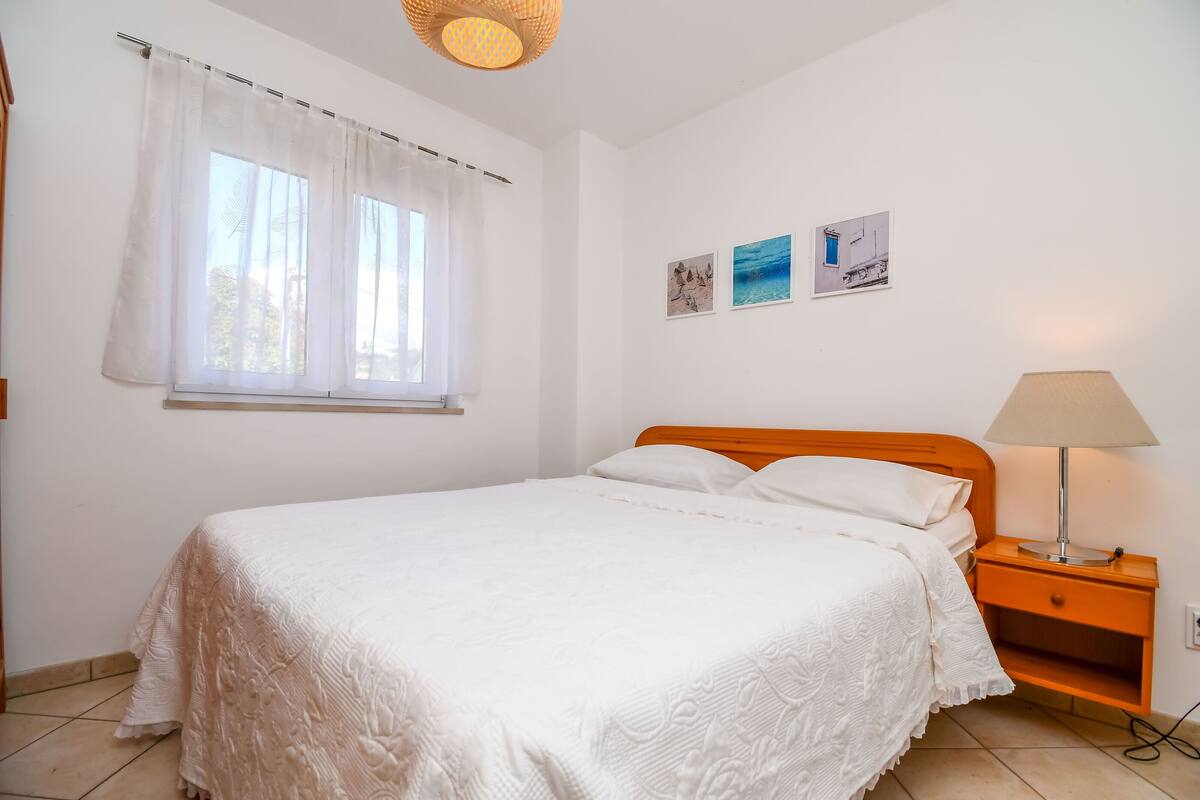 A bedroom with a bed, a nightstand, a lamp, a window with curtains, and two wall art pieces.