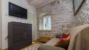 Una habitación con pared de piedra, un televisor de pantalla plana montado sobre un armario de madera oscura, una ventana con contraventanas blancas y un sofá color beige con cojines de colores.