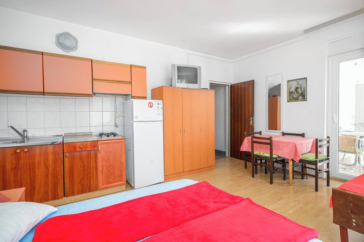A compact kitchen with wooden cabinets, a white refrigerator, and a microwave. There is a dining area with a table and chairs, and a bed with red bedding.
