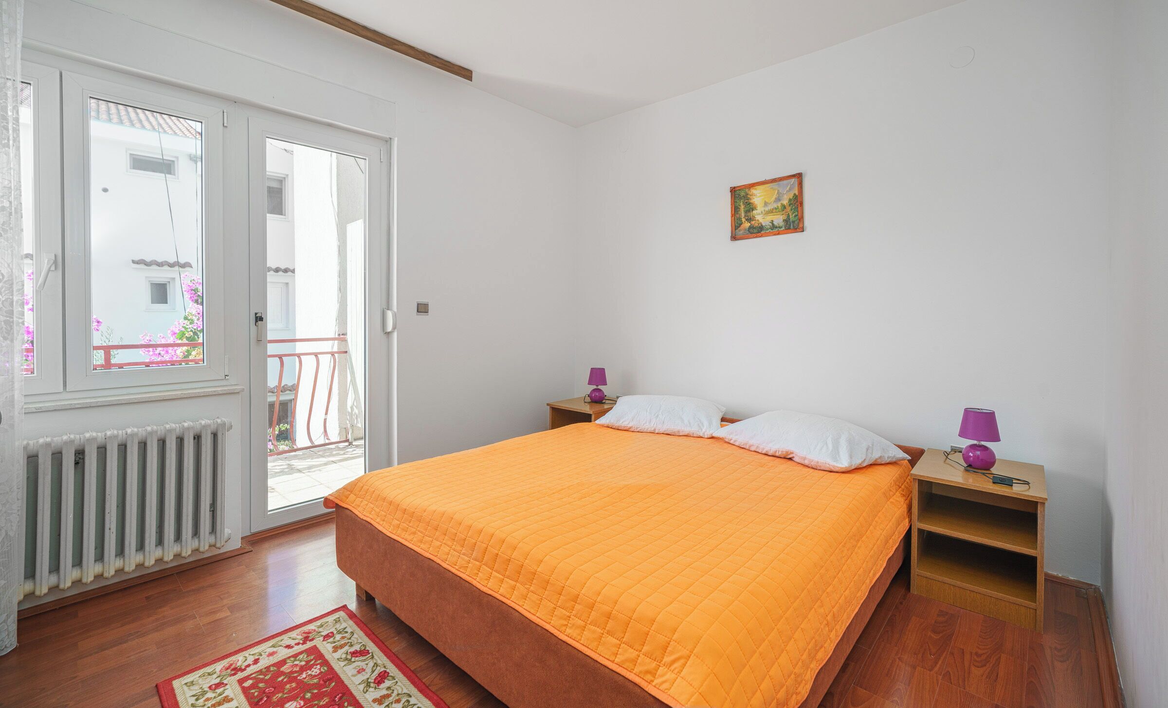 Ein Schlafzimmer mit Bett, Nachttisch, Fenster, Heizkörper und Tür.