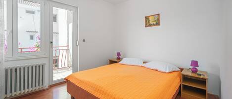 Ein Schlafzimmer mit Bett, Nachttisch, Fenster, Heizkörper und Tür.