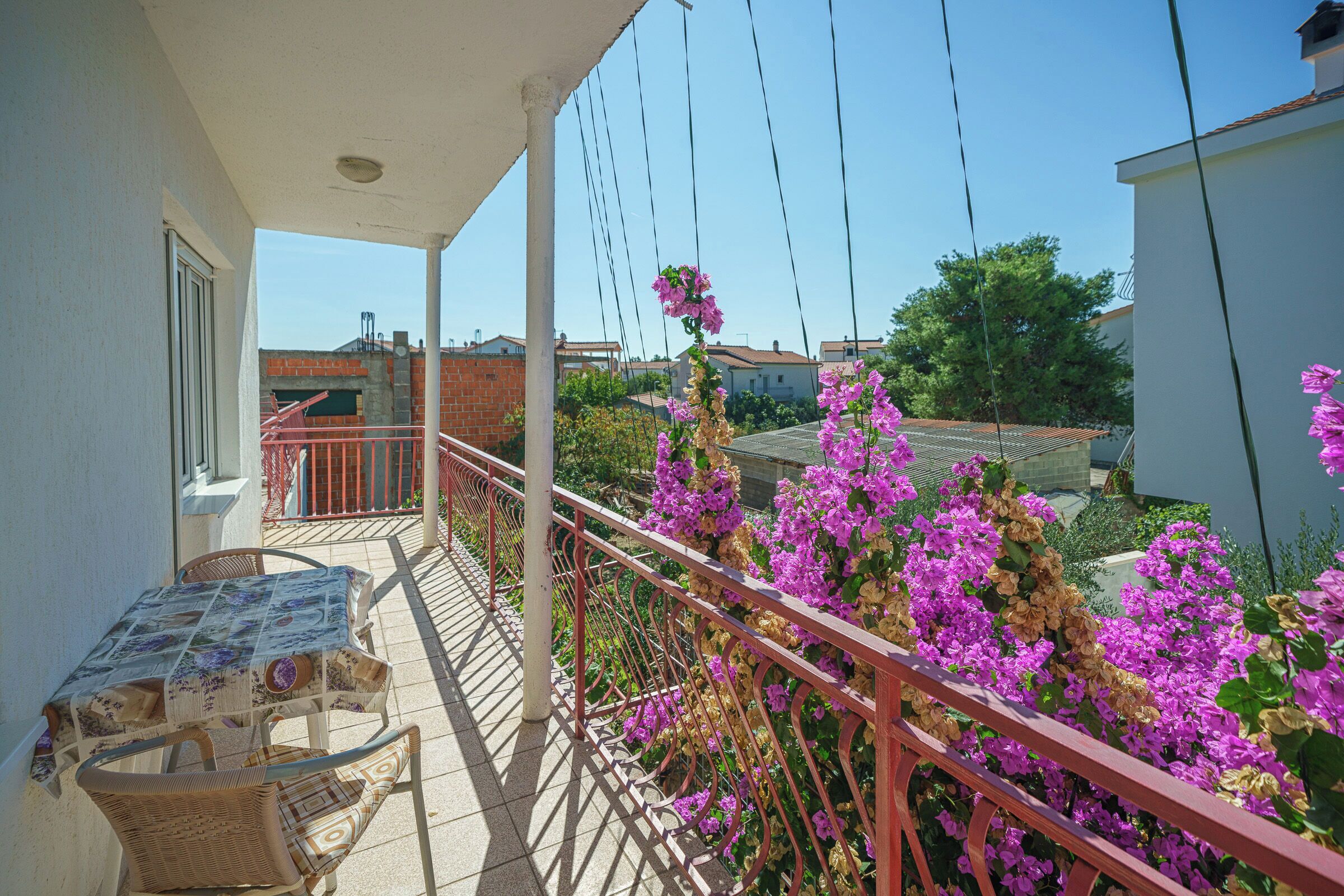 A balcony with a table and chairs, surrounded by a metal railing and blooming flowers.