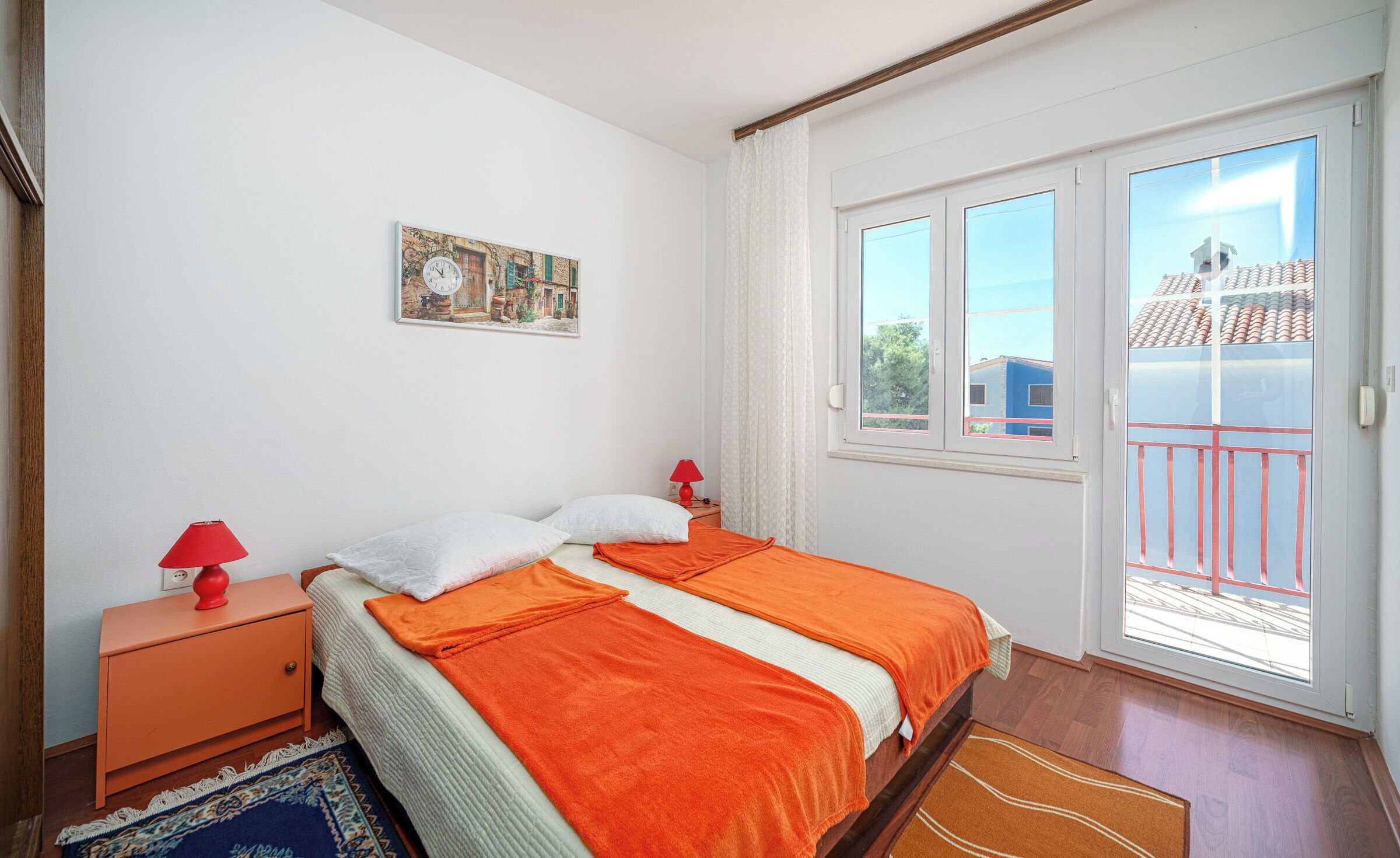A bedroom with a bed, a nightstand, a red lamp, a white pillow, a red lamp on the nightstand, a window with a view of buildings, and a door.