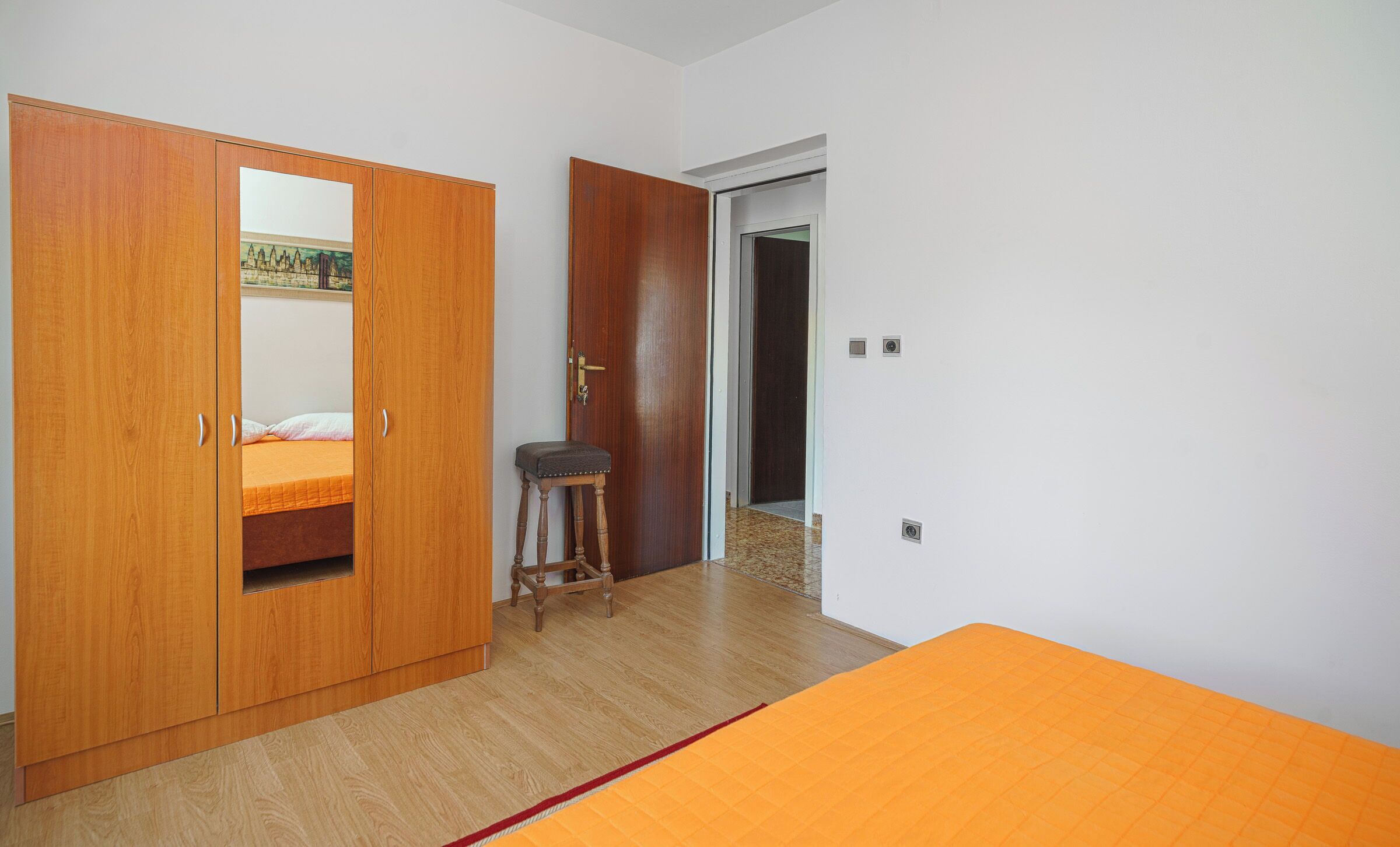 Ein Schlafzimmer mit einem Holzschrank, einem Bett mit orangefarbener Bettdecke und einem kleinen Hocker.