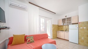 Ein kompakter Wohnraum mit einer Couch, einem kleinen Tisch, einer Küchenzeile mit Schränken und einem Kühlschrank sowie einem an der Wand montierten Fernseher.