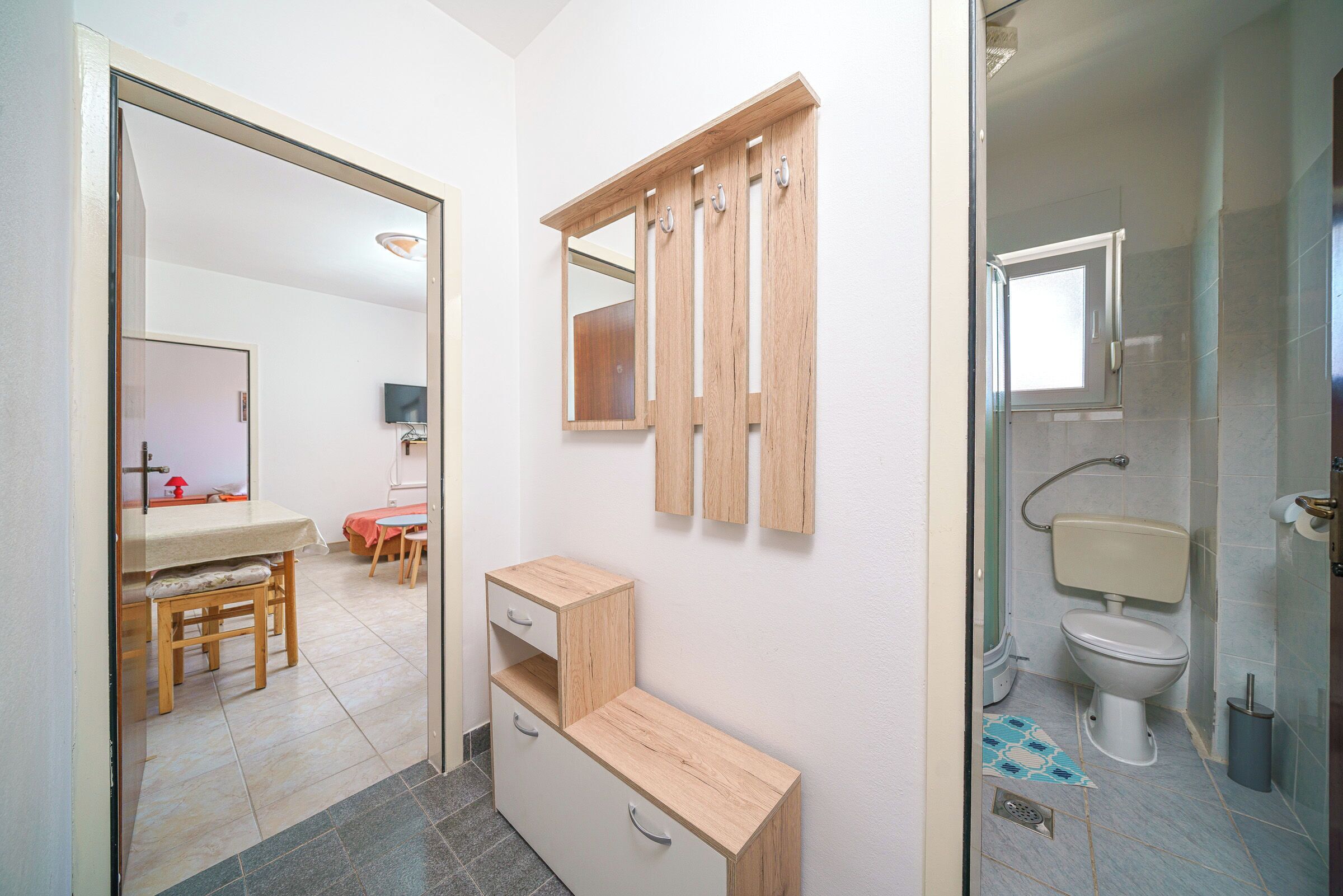 A bathroom with a toilet, sink, and mirror. There is a wooden cabinet with drawers and a small table with chairs in the adjacent room.