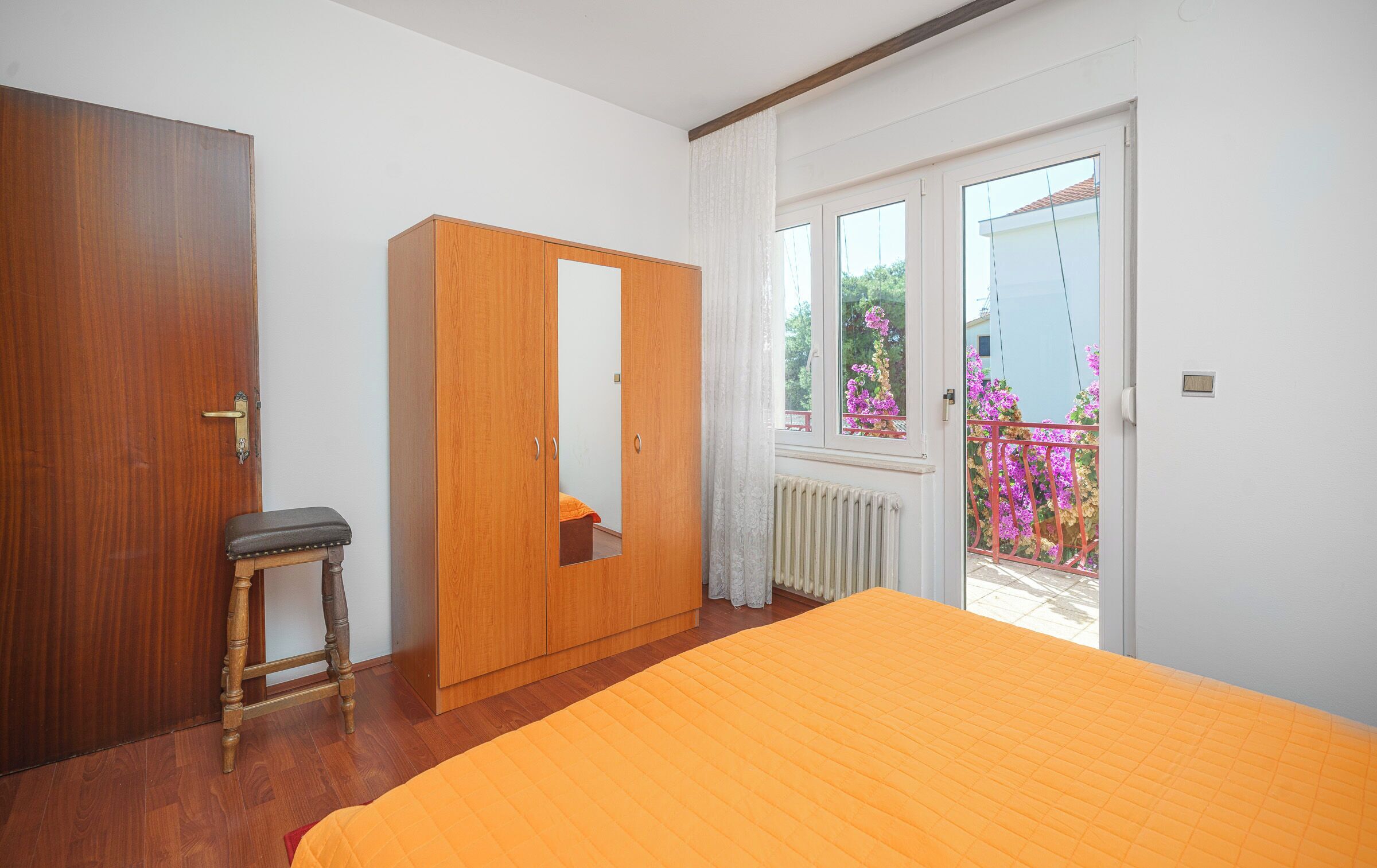 Een slaapkamer met een houten garderobe, een bankje, een bed met oranje sprei en een balkon met bloemen.