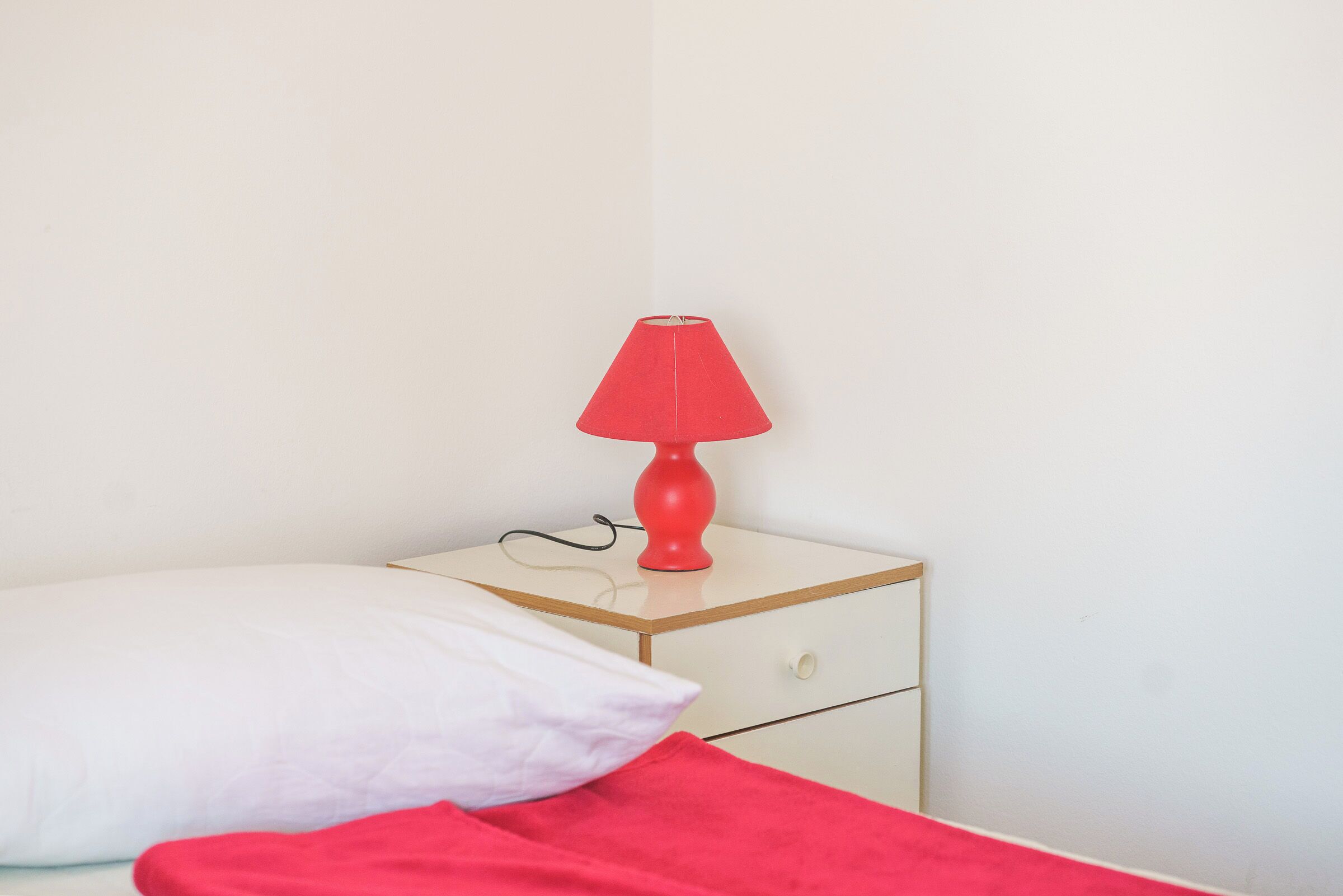 A bed with a white pillow and a red blanket, a red table lamp on a bedside table, and a white wall.