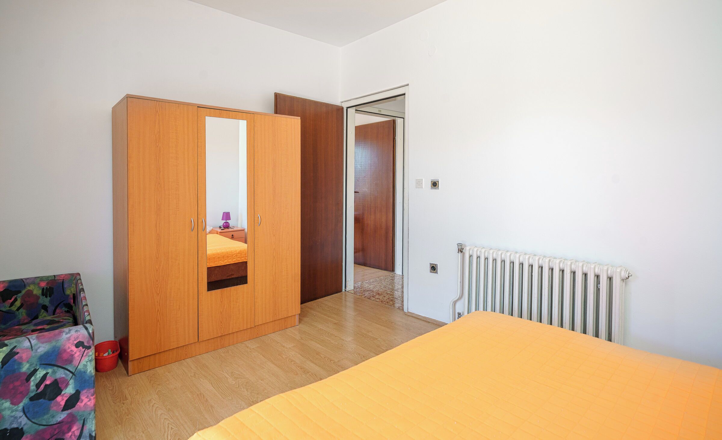Un dormitorio con un armario de madera, una cama con colcha amarilla, un radiador y una silla colorida.