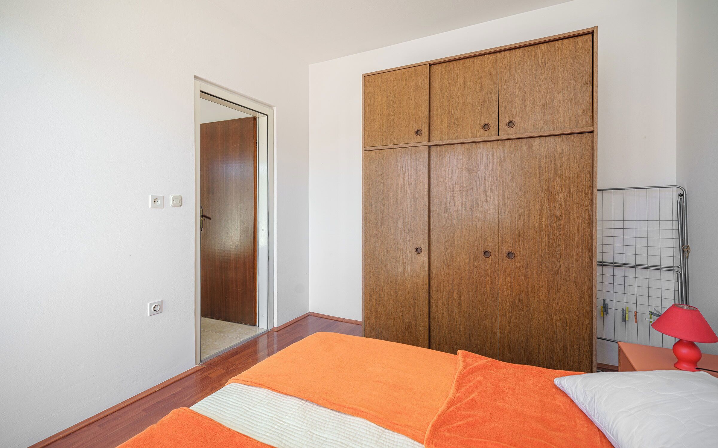 A bedroom with a wooden wardrobe, a bed with orange and white bedding, and a red lamp on a nightstand.
