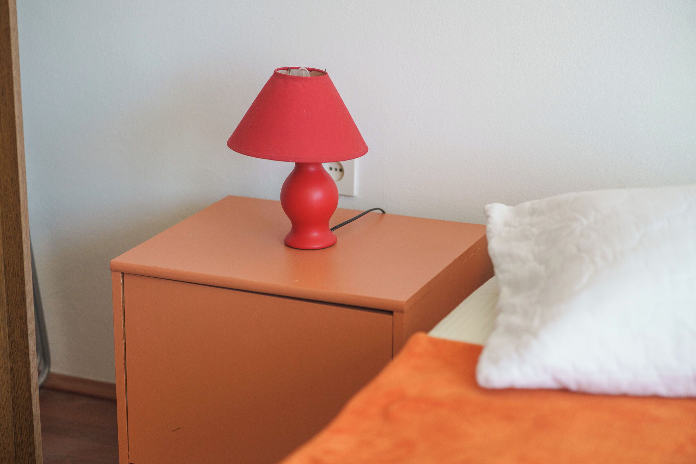 Una mesita de noche naranja pequeña con una lámpara roja y un cojín blanco sobre una cama naranja.
