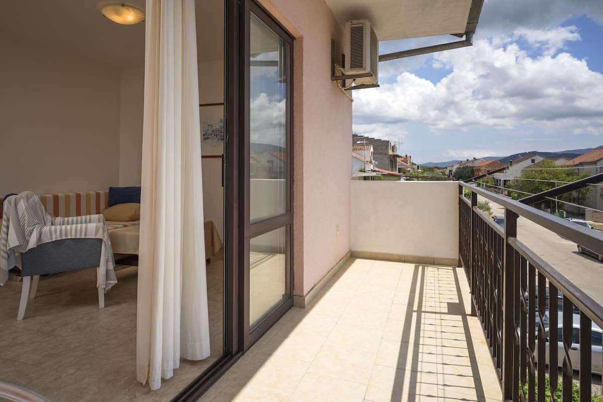 A balcony with a sofa, a table, and a chair, overlooking a residential area with houses and mountains in the background.