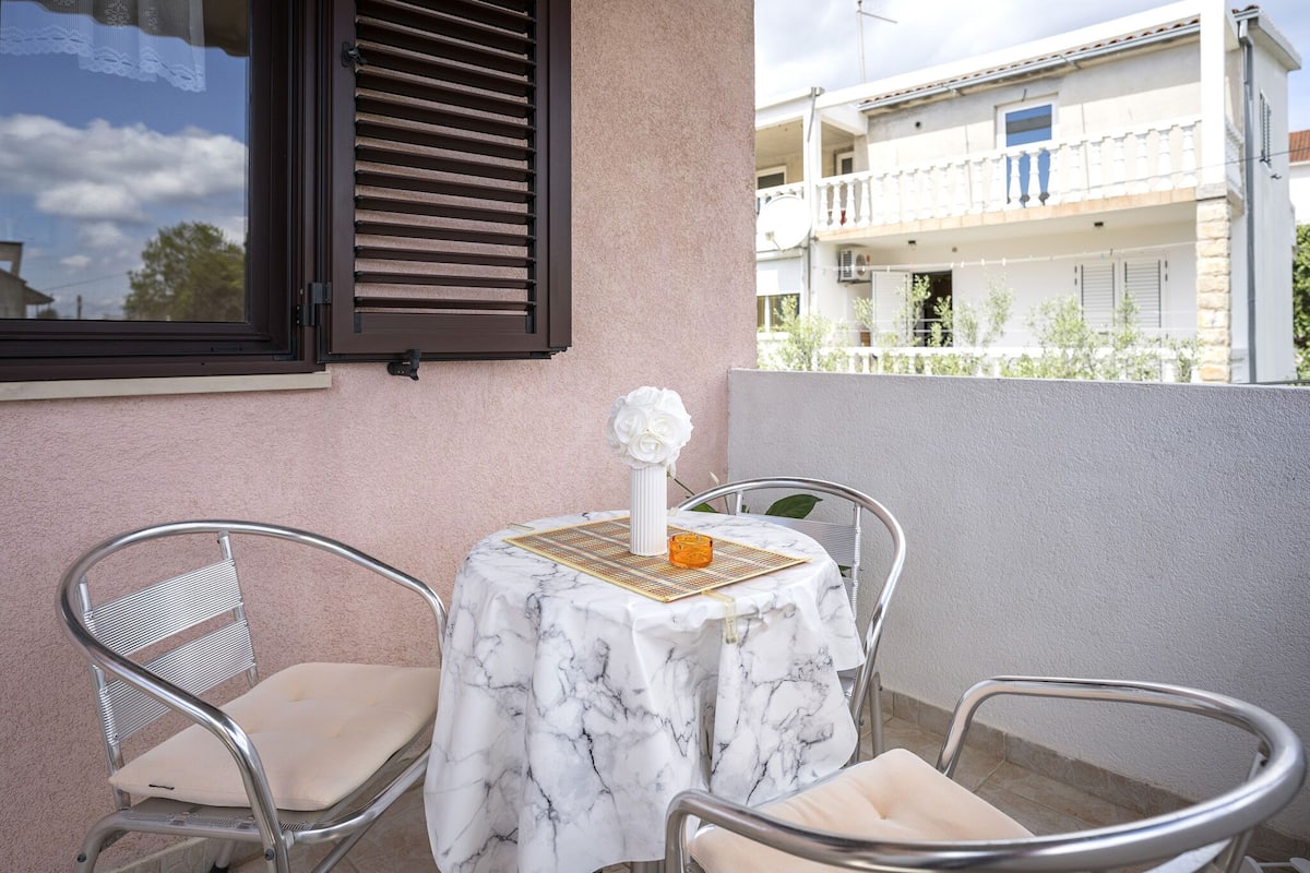 A small outdoor patio with a table covered in a white marble tablecloth, two metal chairs, a decorative vase, and a window with dark shutters.
