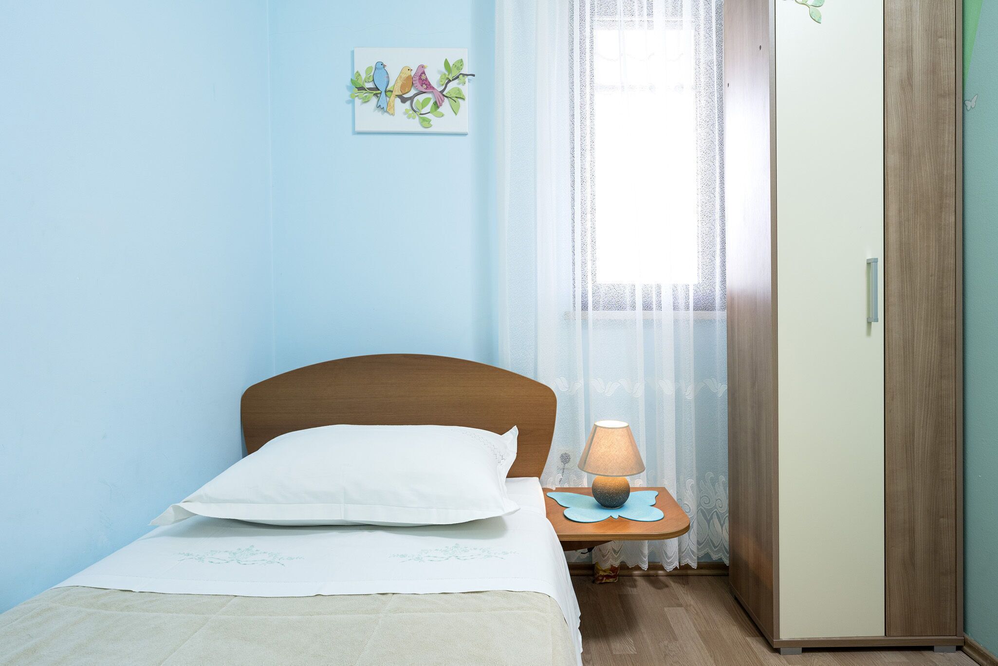 Un dormitorio con cama, mesita de noche con lámpara, armario y ventana con cortinas translúcidas.