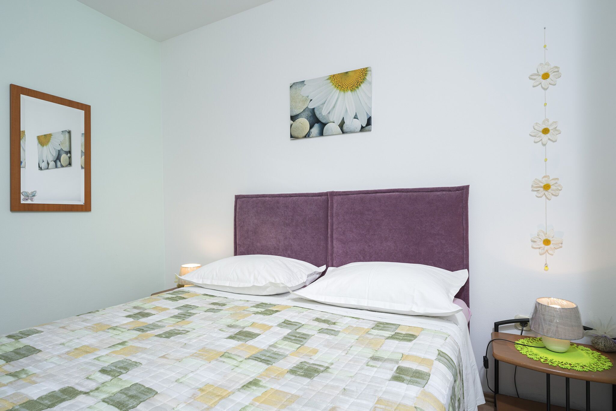 A neatly made bed with a purple headboard, a framed picture of daisies on the wall, and a string of white flowers.